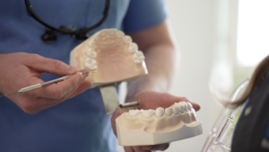 A person in scrubs holds dental models and points at teeth with a dental tool, likely explaining dental care or a procedure to a patient.
