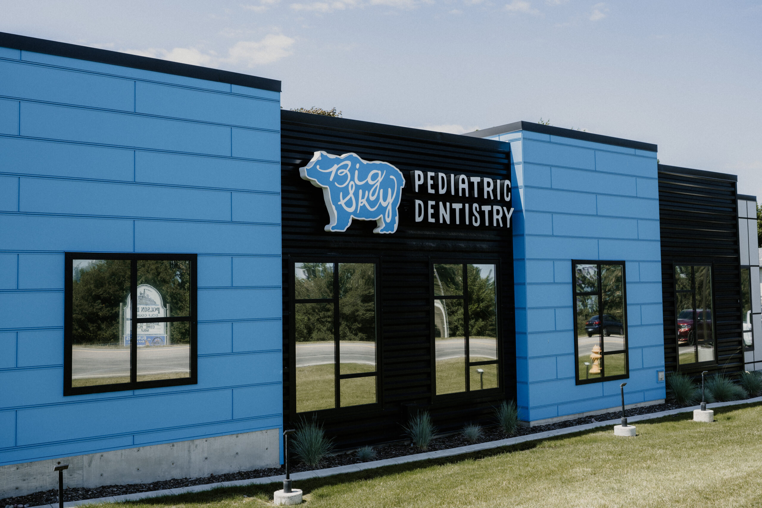 A modern blue and black building with a sign that reads Big Sky Pediatric Dentistry and a bear logo on the front. Green grass and trees are visible in the background.
