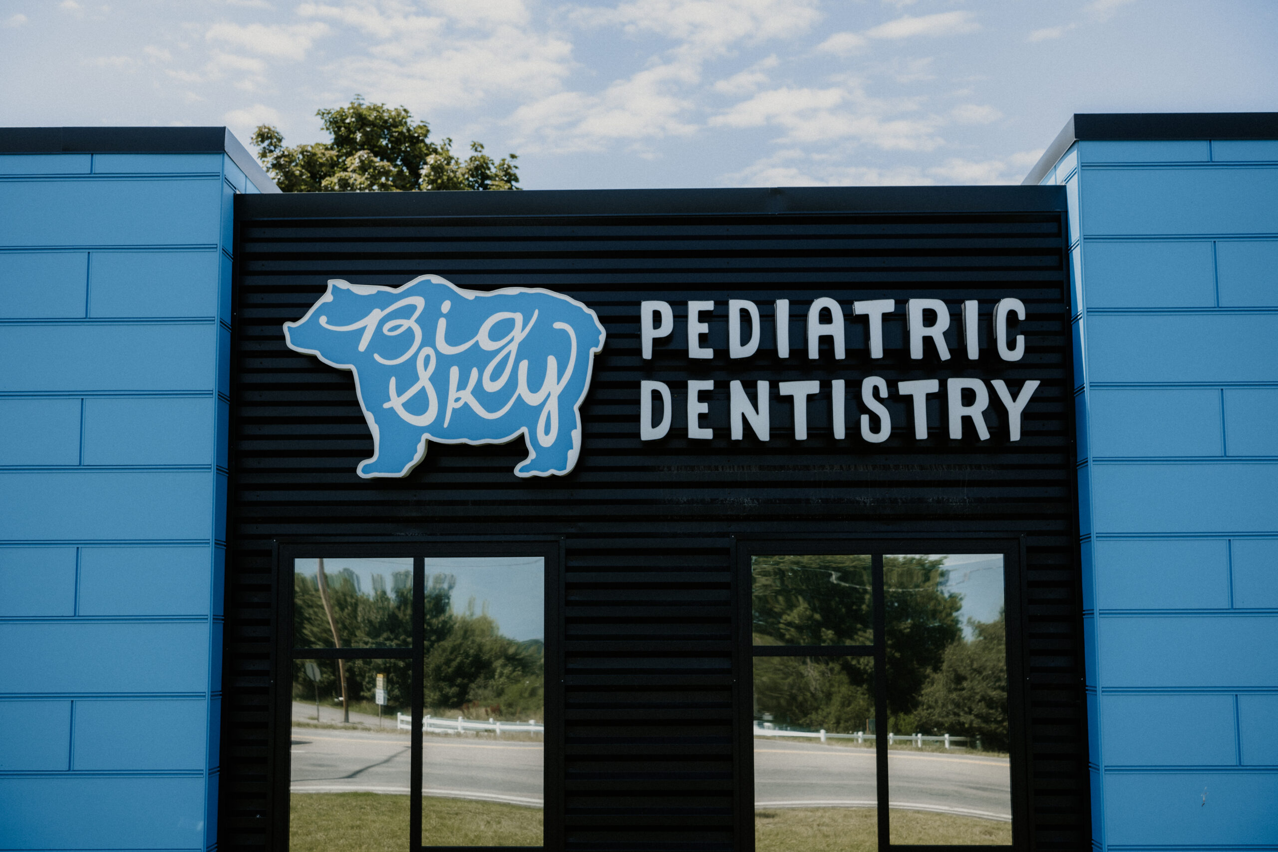 A building with a sign that reads Big Sky Pediatric Dentistry beside a blue bear logo. Blue panels and large windows are visible, with trees and sky reflected in the glass.