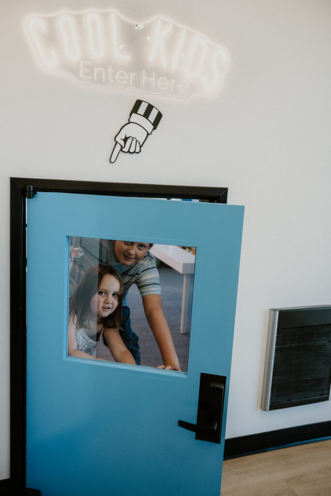 A blue door with a window shows two children, one standing and one sitting. Above the door, a neon sign reads COOL KIDS Enter Here with a pointing hand. The wall is white with a black vent to the right.