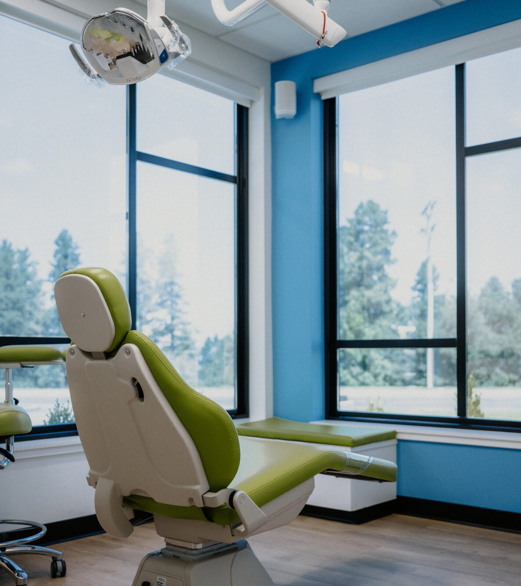 A green dental chair in a bright, modern dental office with large windows, wooden floors, and blue walls. Dental equipment hangs from the ceiling.