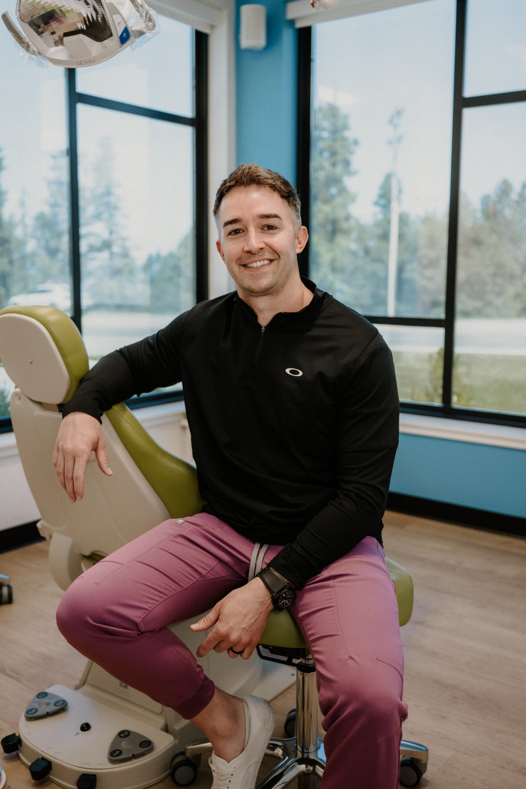 A man in a black shirt and pink pants sits on a green dental chair in a bright room with large windows, smiling at the camera.