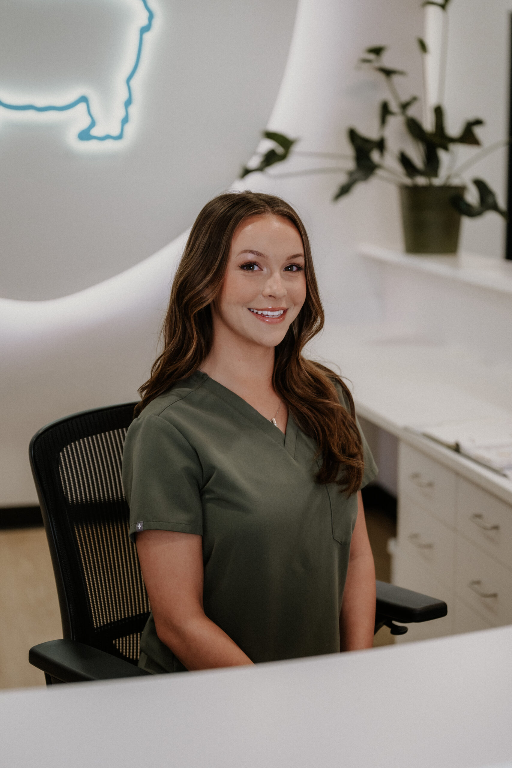 A woman in green scrubs sits and smiles at a reception desk, with plants and papers nearby and a glowing wall decoration behind her.