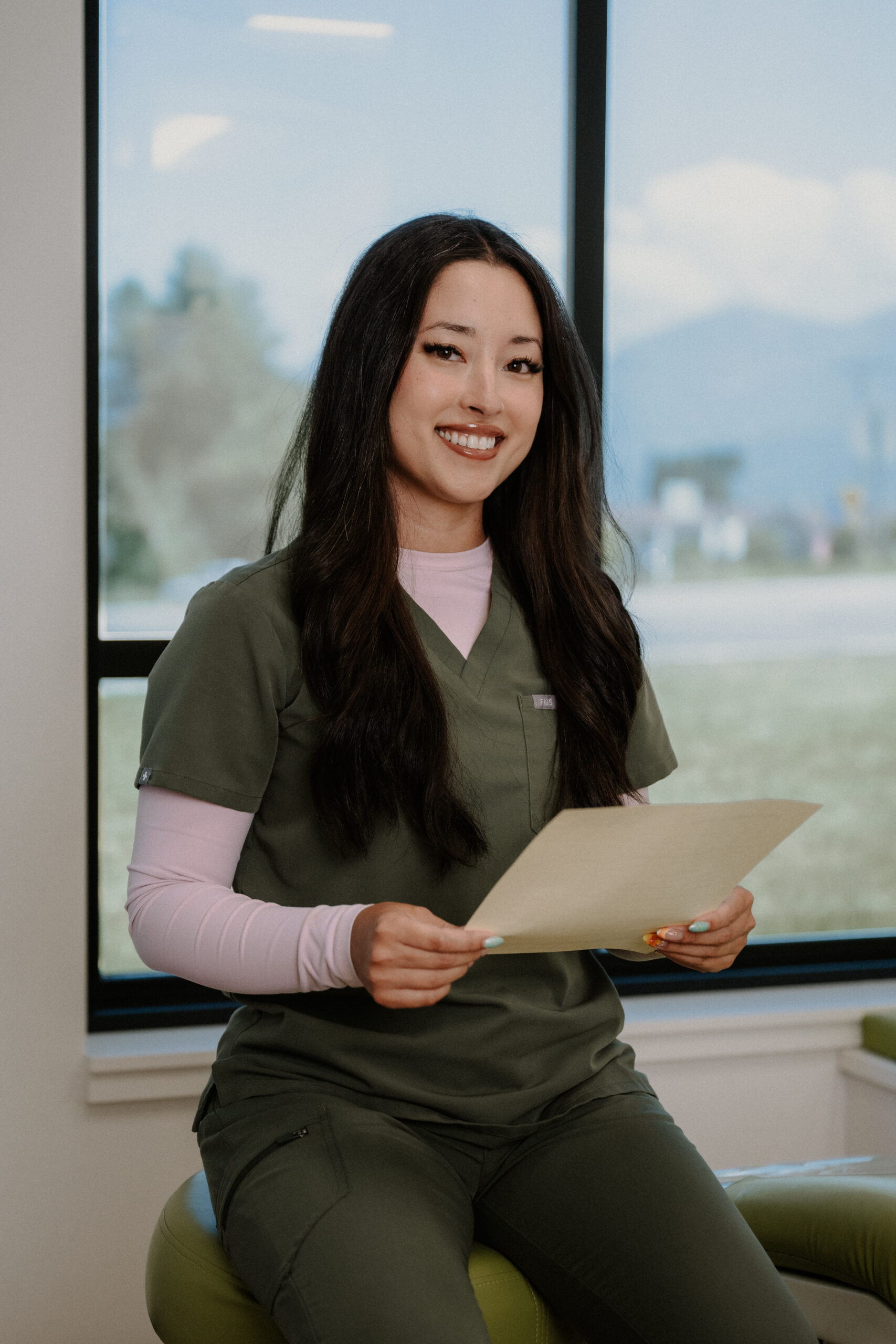 A woman in green scrubs sits indoors by a window, smiling and holding a file folder or document in her hands. She has long dark hair and is seated on a light green stool, with a bright outdoor scene in the background.
