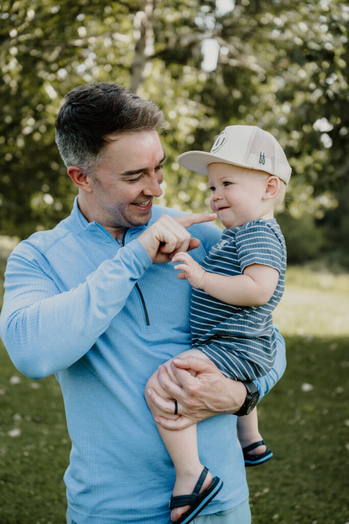 A smiling man in a light blue shirt holds a happy toddler wearing a striped shirt and cap outside in a sunny, green park, gently touching the childs nose.
