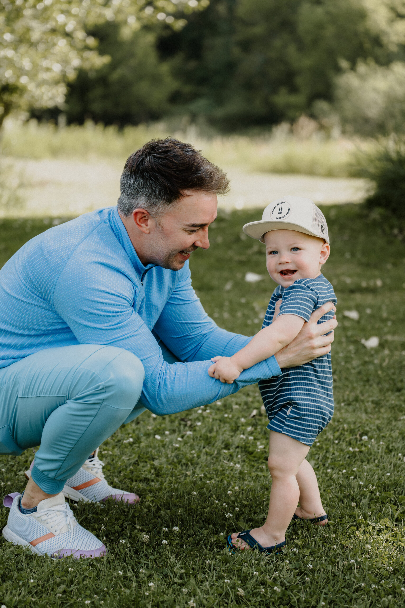 A man in blue holds a smiling toddler wearing a striped romper and cap, outdoors on green grass with trees in the background.