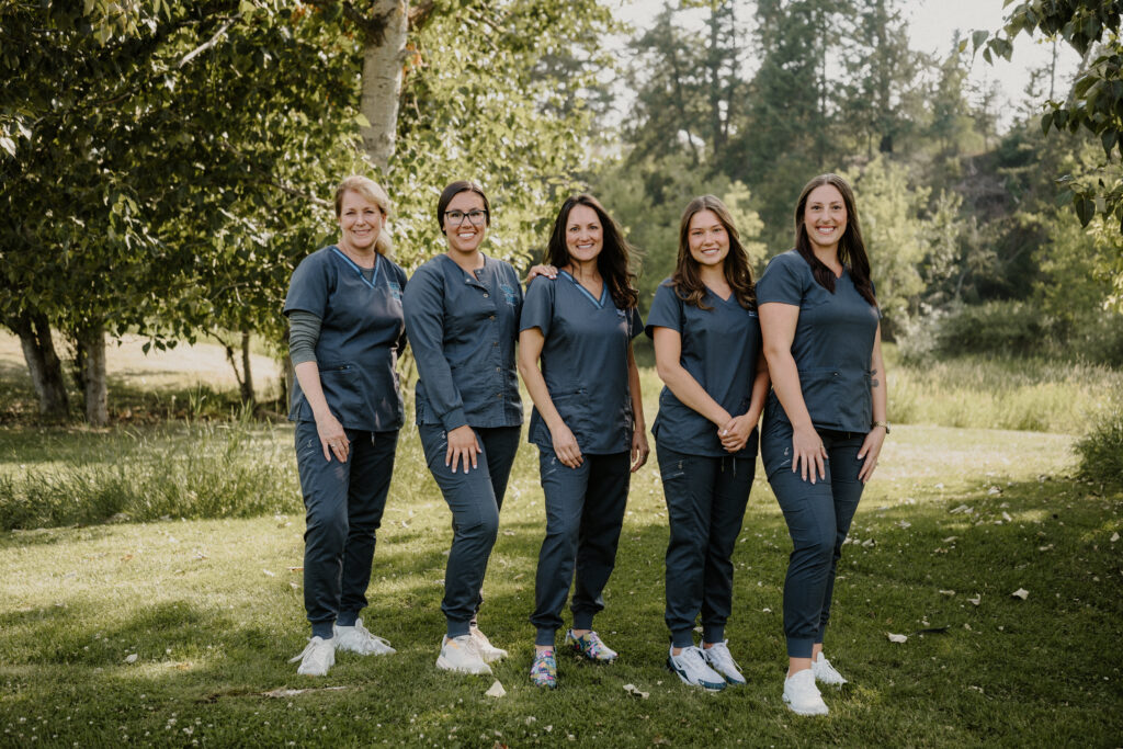 Five women in blue scrubs stand in a line, smiling outdoors on a grassy area with trees and sunlight in the background.