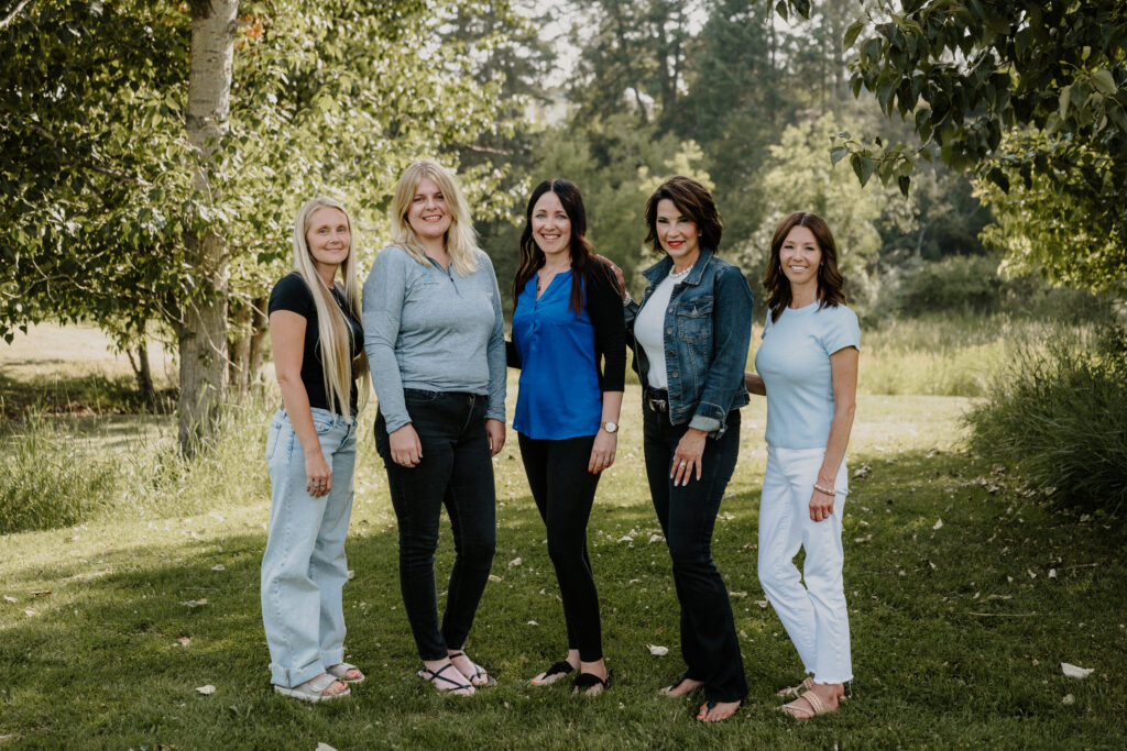 Five women stand outside on grass, smiling, with trees and greenery in the background. They wear casual clothing in light blue, black, and denim tones on a sunny day.