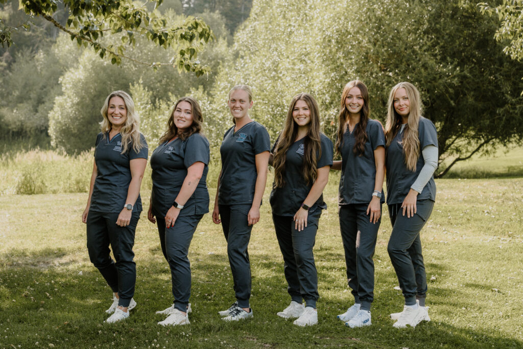 Six women wearing matching dark scrubs and white sneakers stand in a row on grass, smiling outdoors with trees and sunlight in the background.
