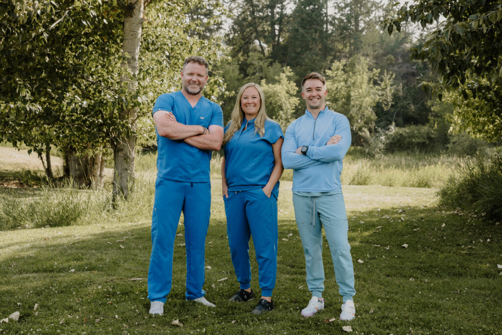 Three people in blue scrubs stand on grass, smiling, with trees and greenery in the background.
