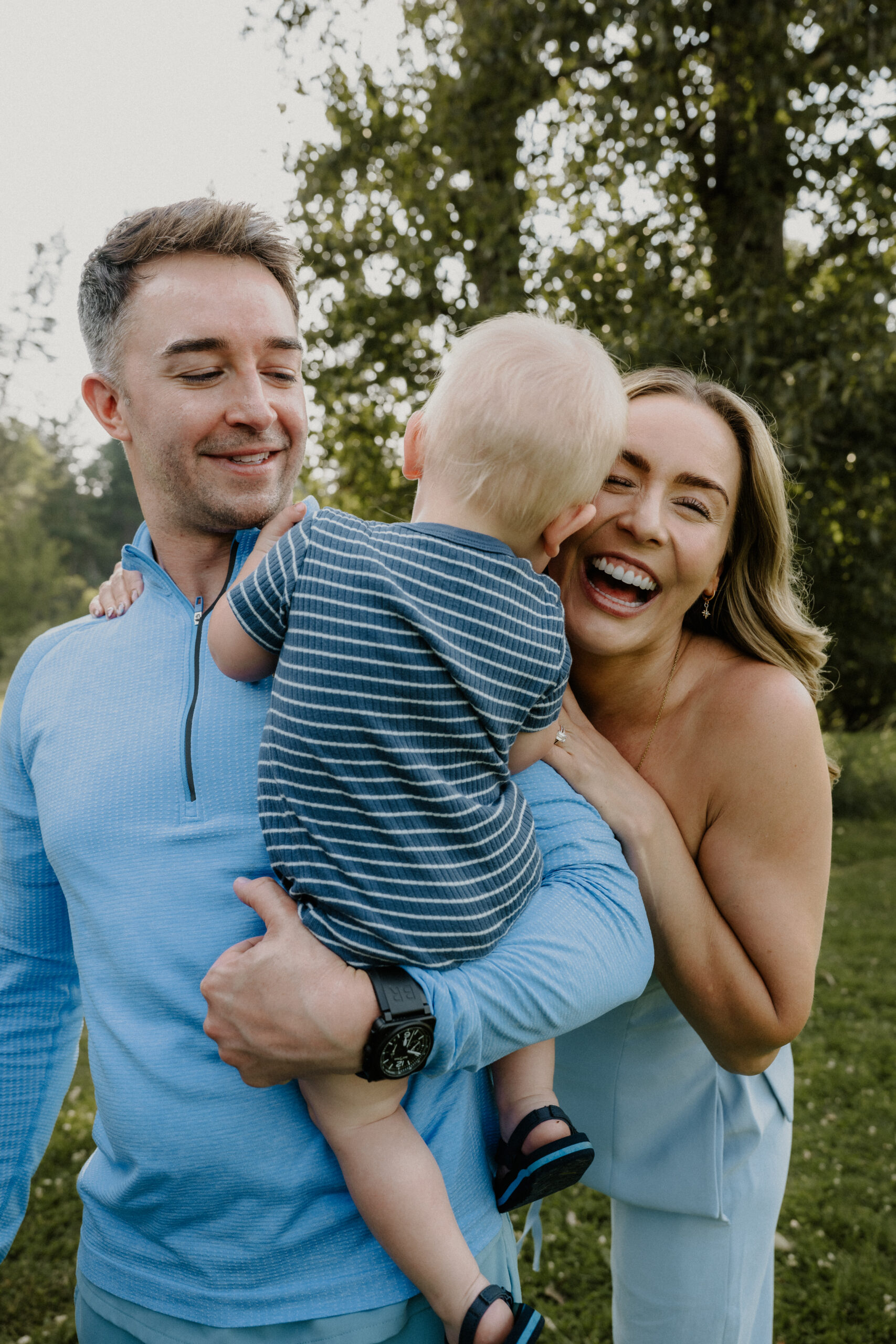 A man holding a small child in striped clothes stands with a smiling woman outdoors. All appear happy, with trees and greenery in the background.